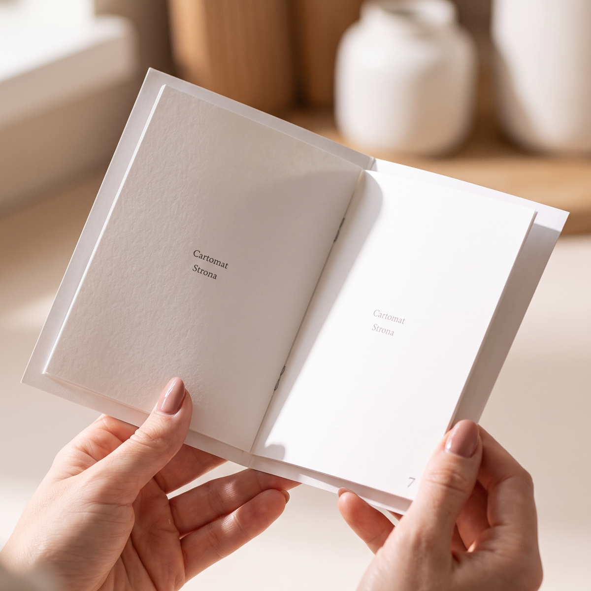 A person holds an open white booklet in both hands, showing blank interior pages. The background is a softly blurred warm indoor setting with wooden decor and white vases.