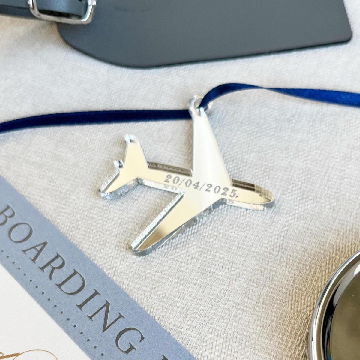 A unique airplane-shaped tag made from mirror silver acrylic, elegantly laser engraved with your wedding date. The tag is strung with a 3mm satin ribbon in blue for a touch of sophistication. It is placed on a textured surface.